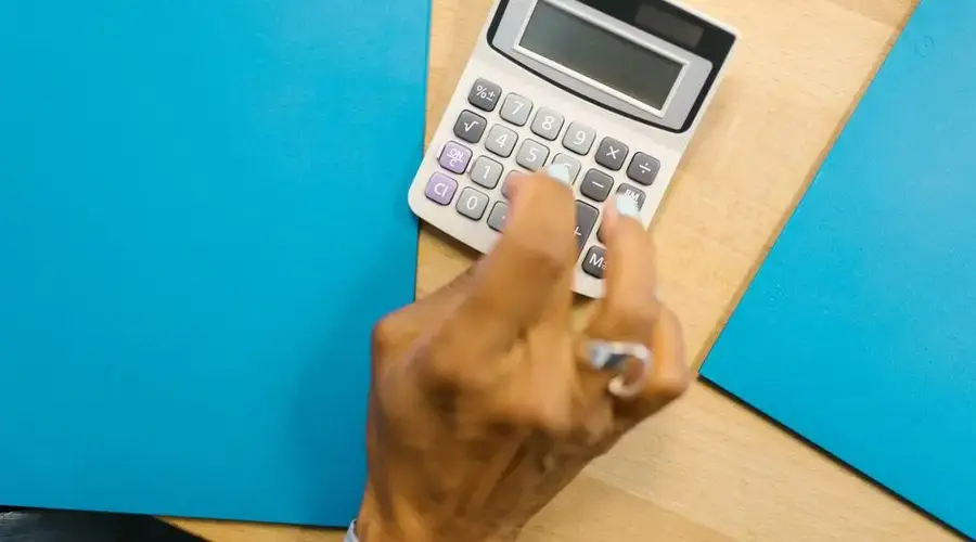 A hand using a calculator on a wooden desk surrounded by blue folders.
