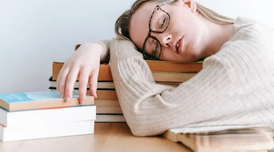 Exhausted female student in eyeglasses and casual sweater sitting at table and sleeping on books on light room
