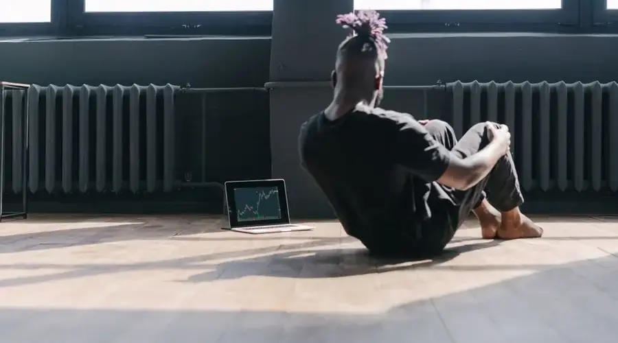 Man doing sit-ups near a laptop showing stock market trends in a bright room.