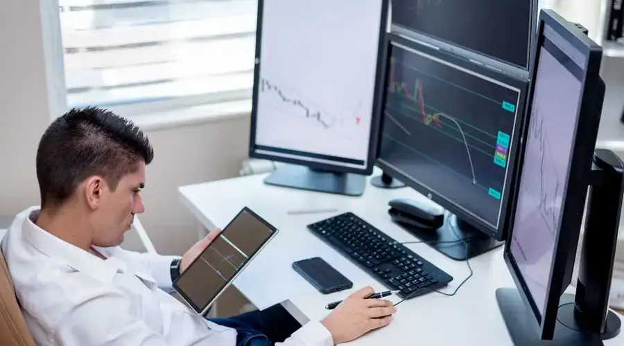 Business professional analyzing financial charts on monitors and tablet in modern office workspace.