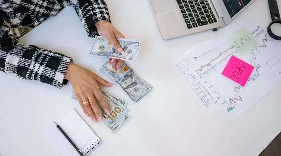 Businesswoman counting dollar bills with financial charts and laptop on table. Investment and finance concept.
