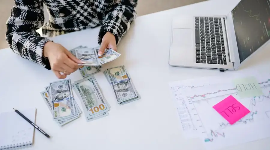 Person counting cash next to laptop and stock market charts on a white table.