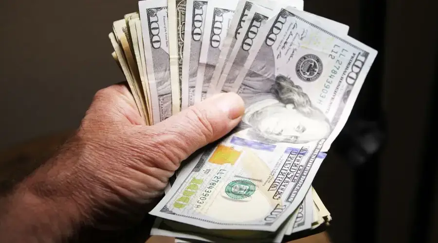 Close-up of a hand holding multiple hundred dollar bills on a dark background.