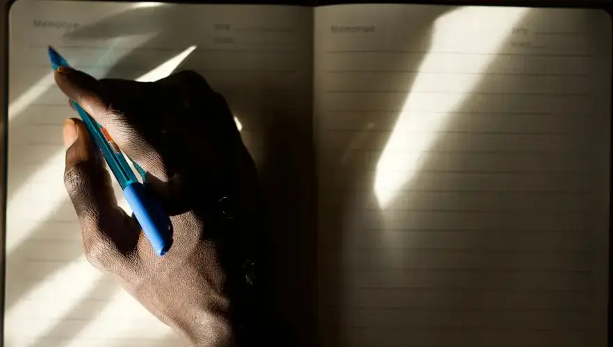 An individual writes notes in a lined notebook, captured in dramatic light and shadow.
