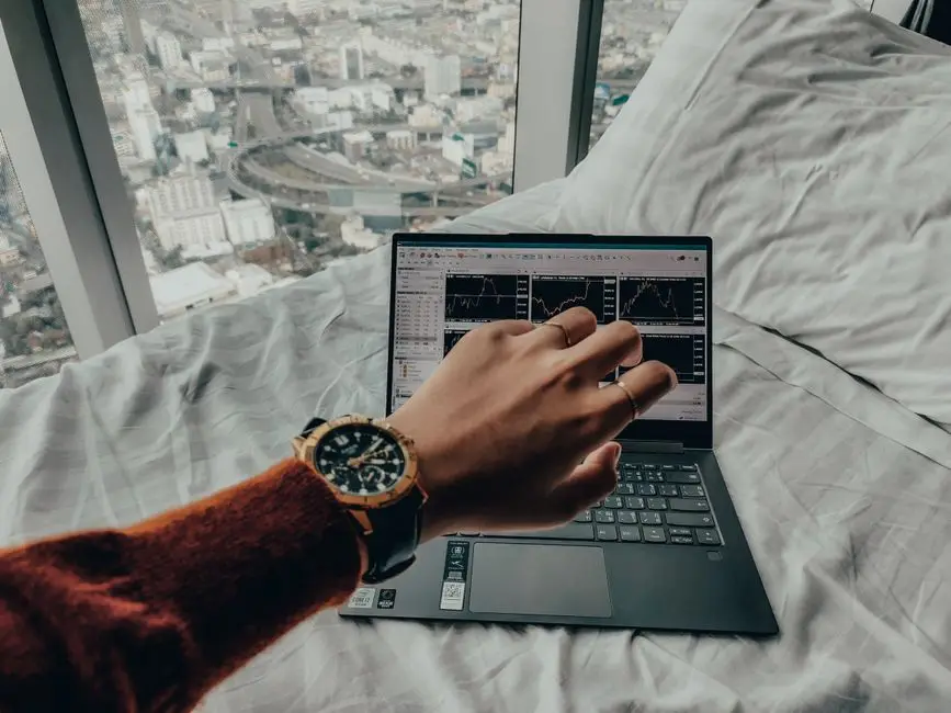 A person checks time while trading on a laptop with charts, overlooking a city view from bed.