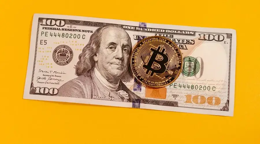 Gold bitcoin placed on a 100 US dollar bill over a bright yellow backdrop, symbolizing modern digital currency.