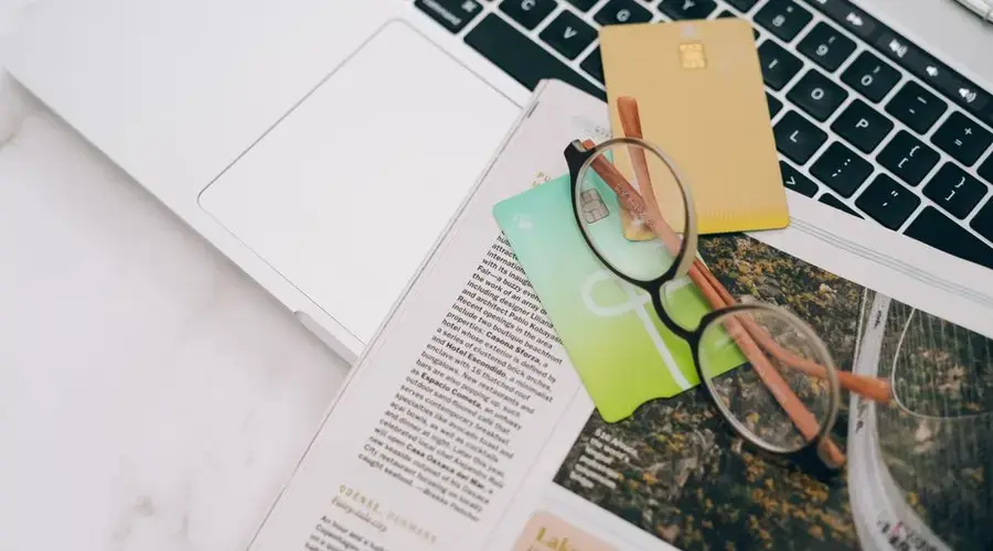 Eyeglasses resting on a magazine beside a laptop and credit cards, indoors.