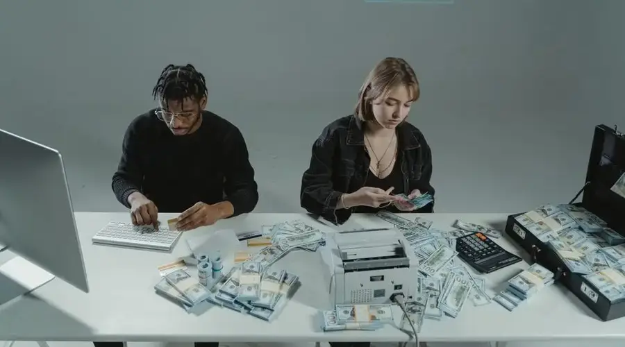 Two people counting money at a desk with computers and cash equipment.