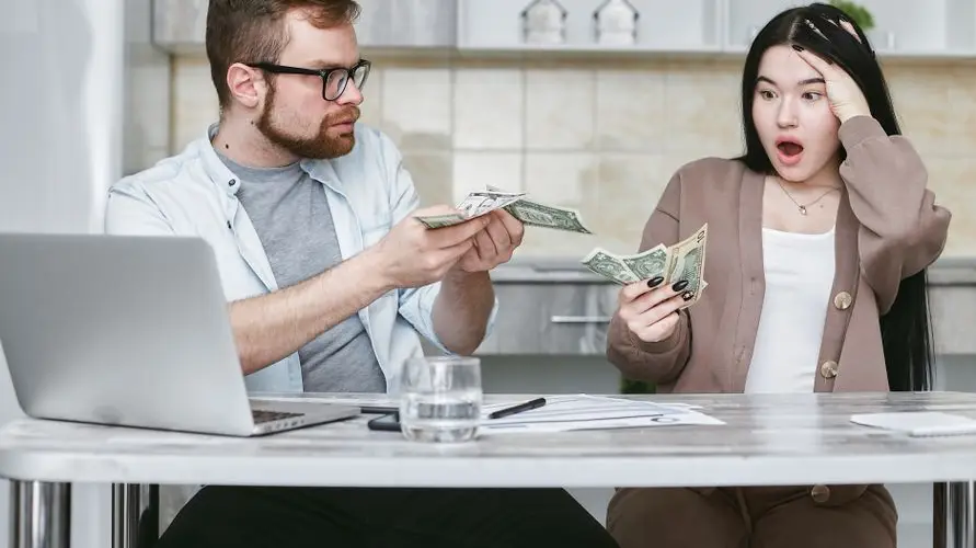 A shocked couple sitting at a table counting cash, illustrating financial surprise.