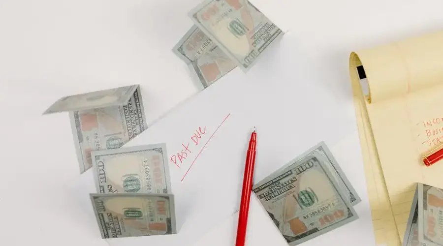 A top-down view of scattered US dollar bills with a 'past due' envelope, red pen, and notepad.