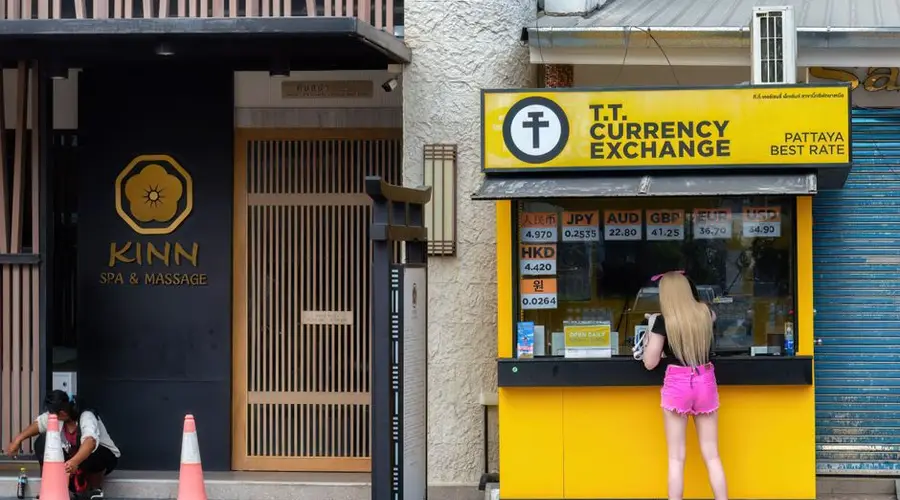 Woman at currency exchange booth in Pattaya, Thailand, with currency rates displayed.