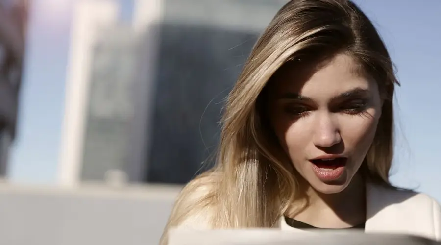 A young woman with blonde hair expresses surprise while reading a newspaper outdoors in daylight.