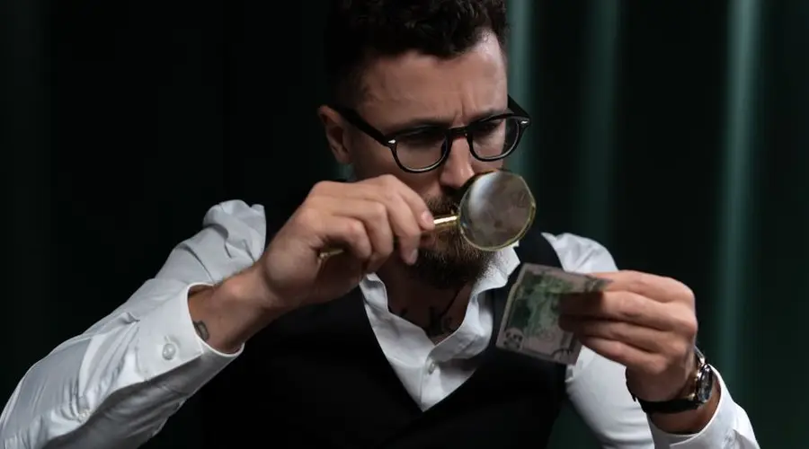 Focused adult inspecting a banknote closely with a magnifying glass indoors.