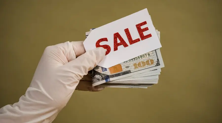 A hand wearing latex gloves holds a stack of dollar bills with a sale sign, symbolizing discount or cash transaction.