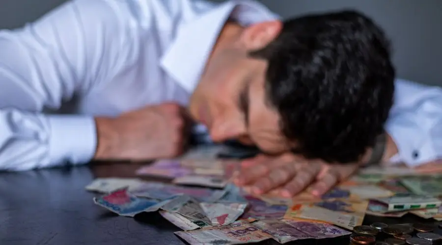 A man in a white shirt rests his head on a table covered with Argentine pesos and coins.