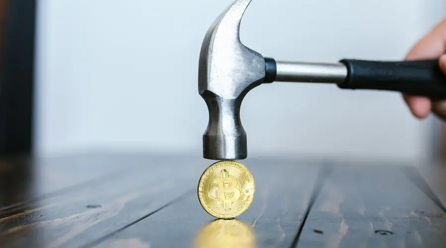 Close-up of a hammer poised above a Bitcoin coin on a wooden surface, representing cryptocurrency challenges.