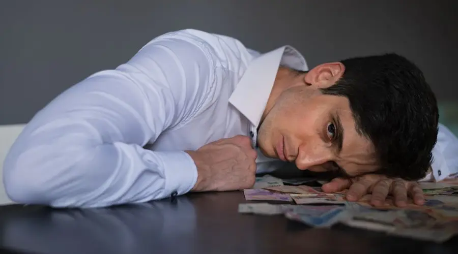Adult man in shirt lying on table, surrounded by currency, looking stressed indoors.