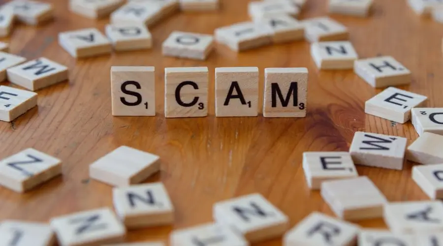 Image of Scrabble tiles spelling 'scam' surrounded by scattered letters on a wooden table.