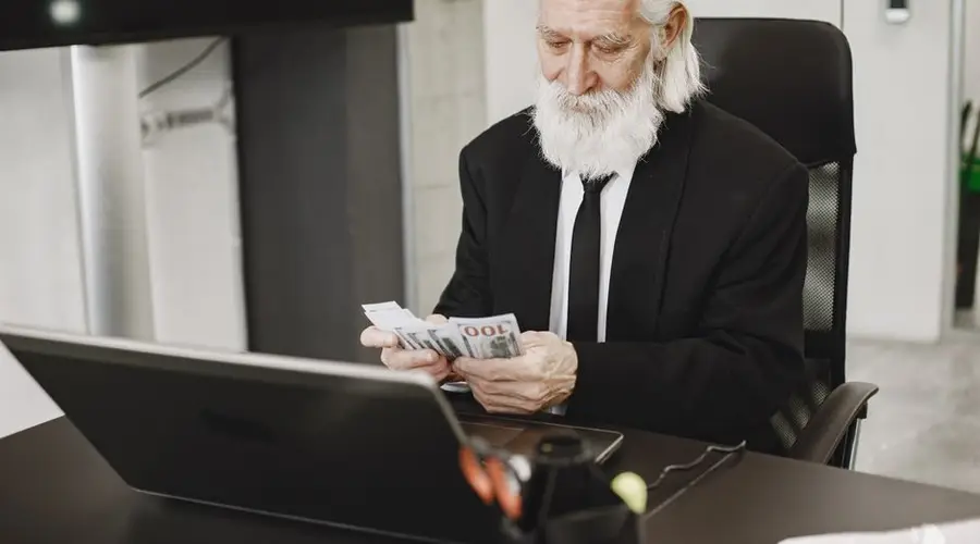 Senior businessman in suit counting money at office desk with a laptop. Elegance and financial focus in modern workspace.