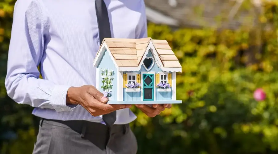 Businessman holds small house model outdoors, symbolizing real estate investment.
