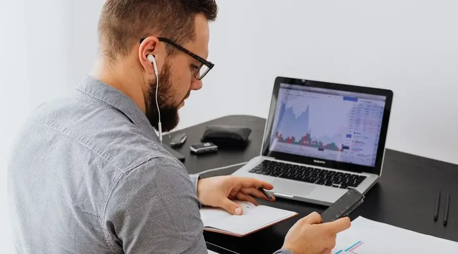 A businessman with earphones analyzes stock market graphs on a laptop while using a smartphone and notebook.