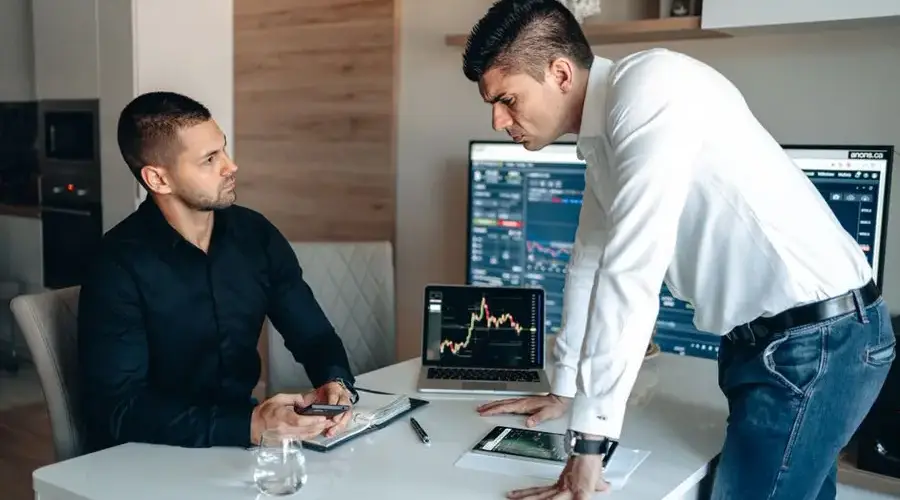 Two businessmen analyzing stock market data on laptops and tablets in an office environment.