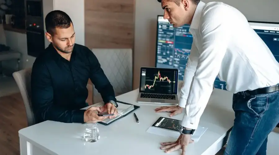 Two businessmen discussing digital market trends with laptops and tablets in a modern office.