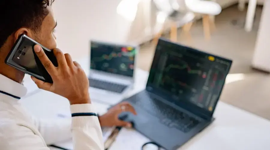 A business professional using a laptop and phone for stock market analysis in a modern workspace.