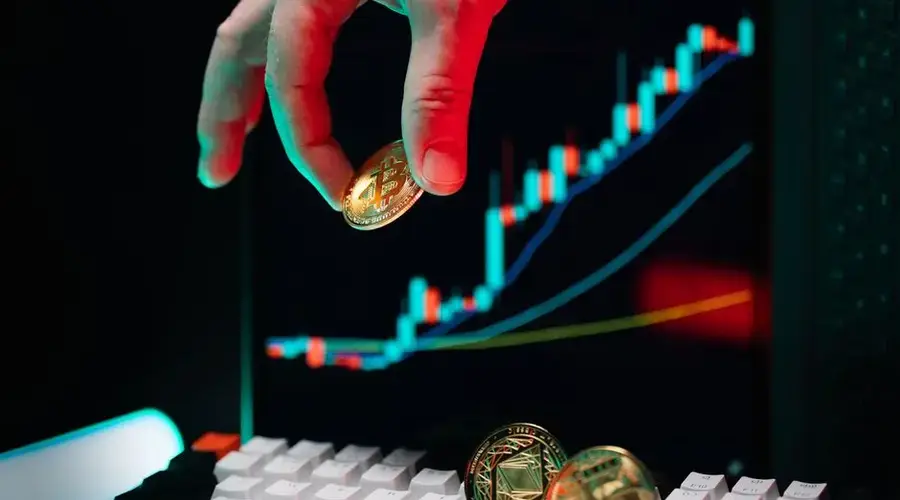 Hand holding Bitcoin with an upward cryptocurrency market chart on screen displaying growth.