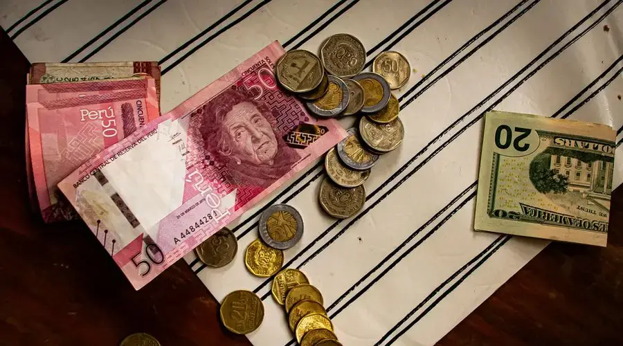 Image of Peruvian banknotes and US dollar bills with scattered coins on a striped surface.