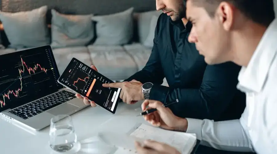 Two business professionals using a laptop and tablet to analyze online trading data indoors.