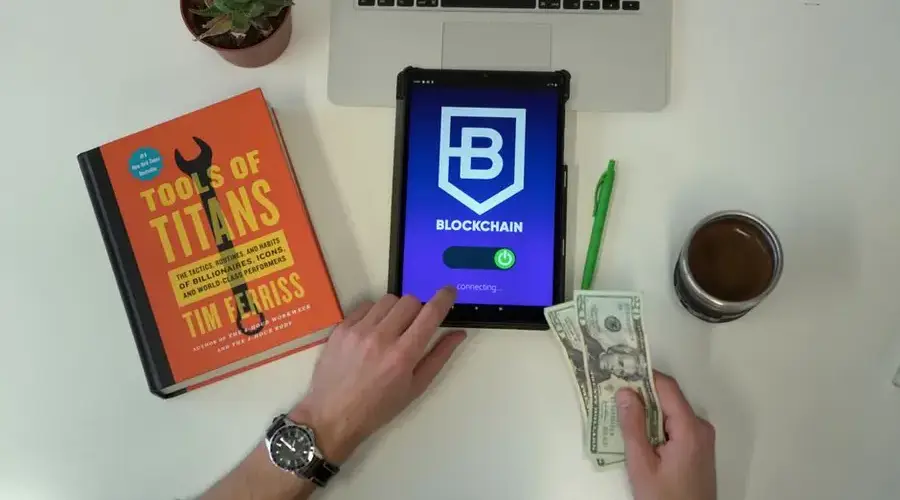 Flat lay image featuring a tablet with blockchain app, US dollars, and a book on a desk.