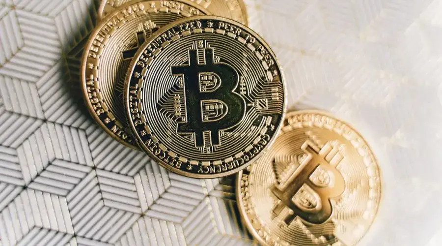 A close-up image of gold Bitcoin cryptocurrency coins on a geometric patterned surface.