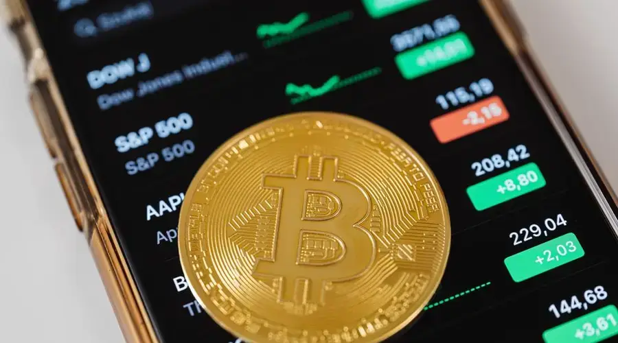 A close-up of a Bitcoin coin placed on a mobile device displaying stock market trading data.
