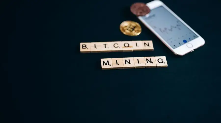 Scrabble tiles spelling Bitcoin Mining with smartphone and coins on a dark background.