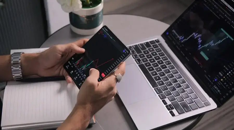 Man analyzing stock trends with laptop and mobile trading app at a modern workspace.