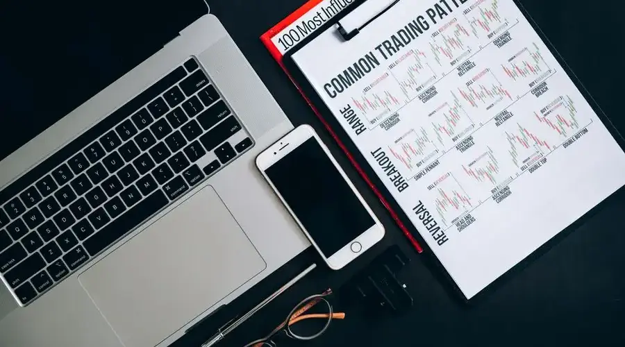 Close-up of office tools including a laptop, smartphone, and trading patterns document on a dark surface.