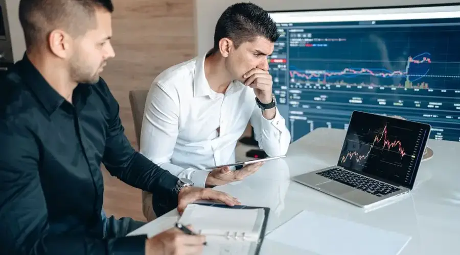 Two businessmen analyzing stock market data on laptops and tablets in an office setting.