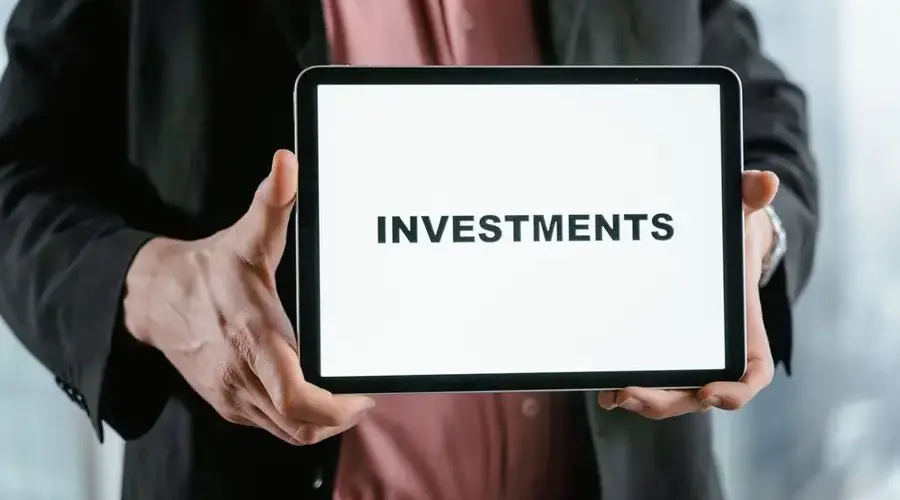 A businessman in a suit holds a tablet displaying the text 'INVESTMENTS'.