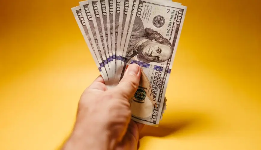 Close-up of a hand holding US hundred dollar bills against a vibrant yellow background.