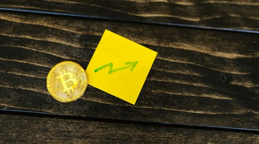 Close-up of a Bitcoin coin with an upward arrow sticky note on a wooden surface.