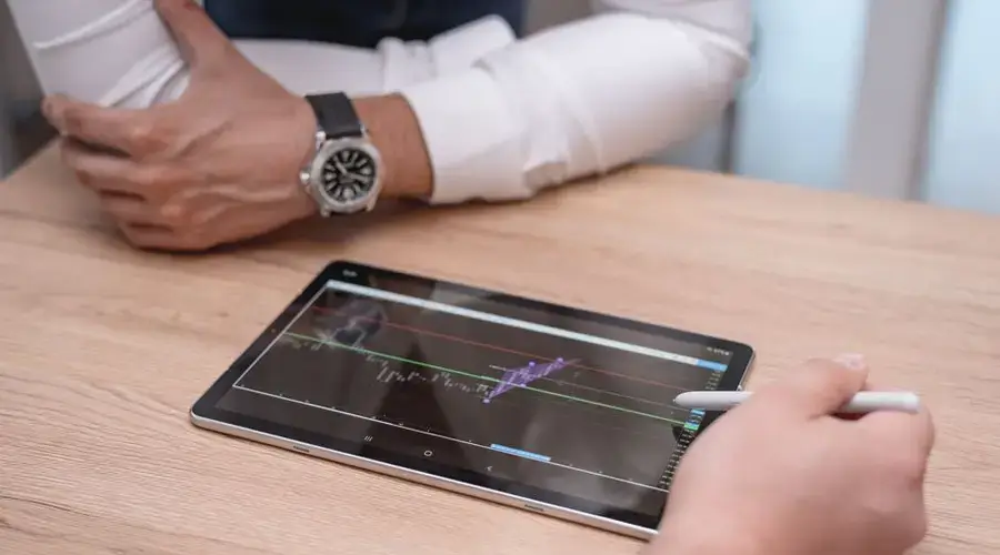 Close-up of hands using a digital tablet with trading charts on a wooden desk.