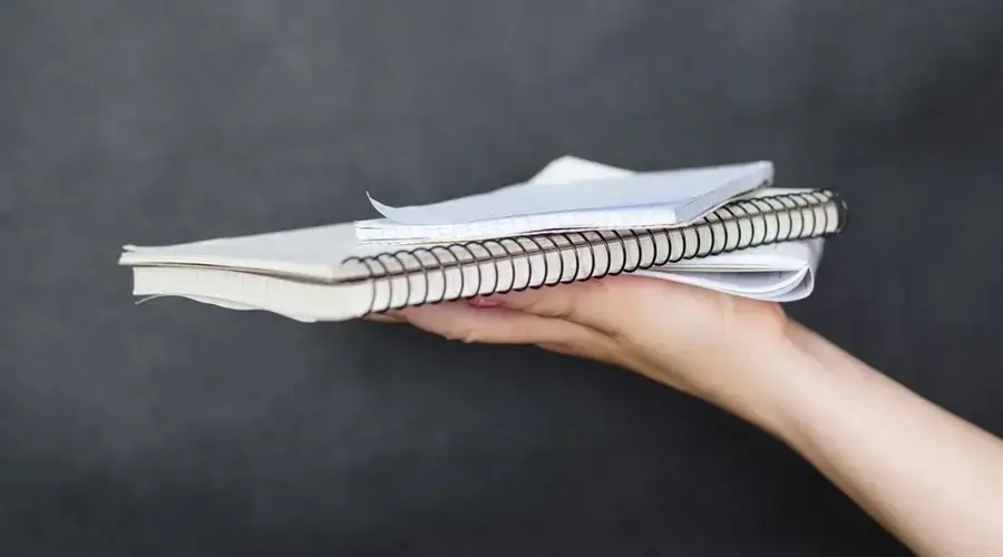 Close-up image of a woman's hand holding a stack of spiral-bound notebooks and papers against a dark background.