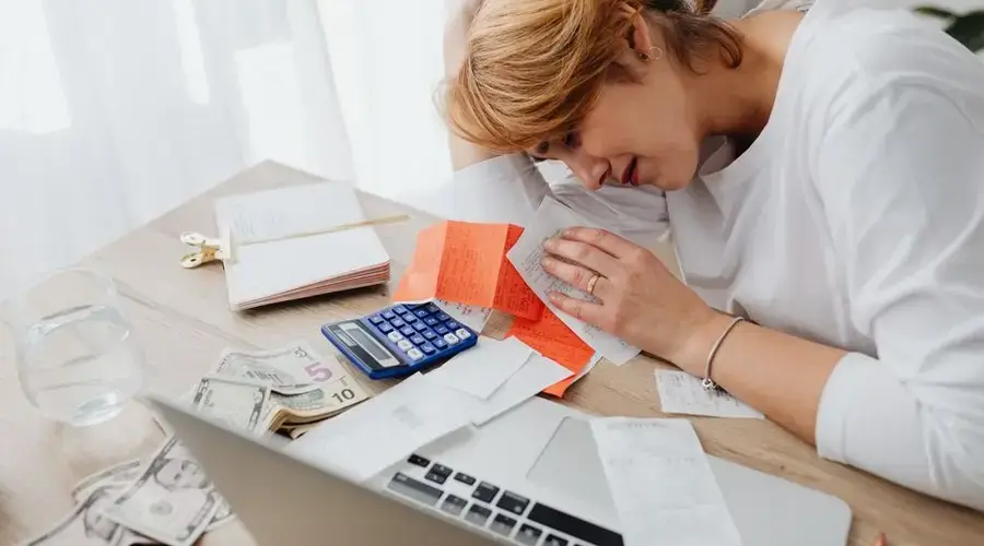 A woman overwhelmed with finances, surrounded by bills, calculator, and cash at home.