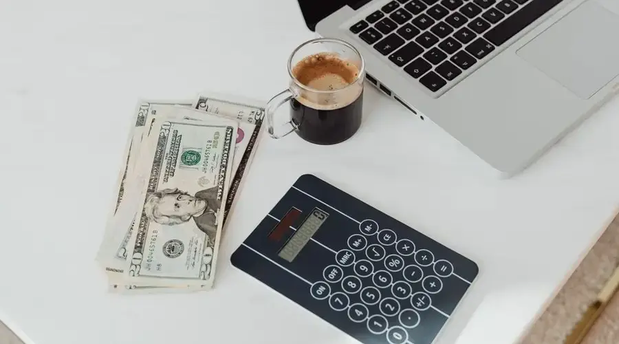 A modern workspace featuring a calculator, cash, and laptop with coffee.