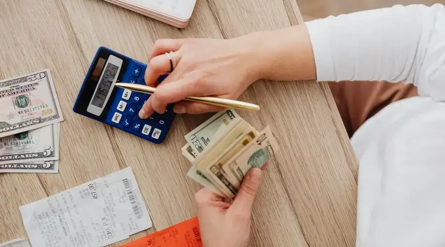 Person using a calculator and counting cash on a wooden table with receipts.