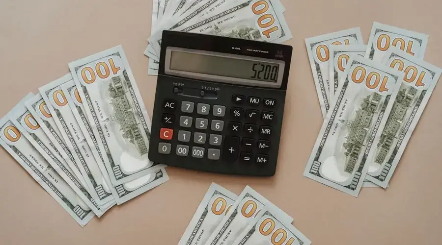Top view of a calculator showing $5200 surrounded by $100 bills on a tan background.