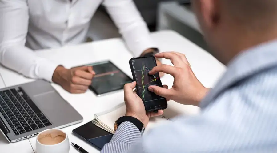 Business professionals using smartphones and tablets to analyze trading charts during a meeting.