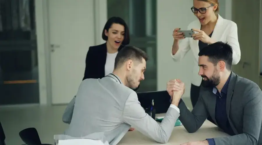 Two businessmen arm wrestling in an office with colleagues cheering them on.