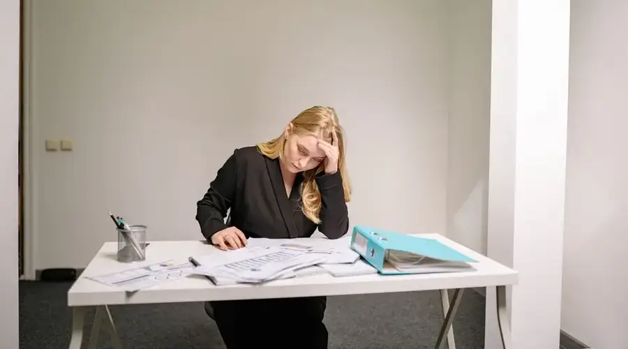 A stressed businesswoman reviewing documents at her desk, overwhelmed by paperwork.